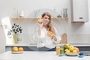 A young woman holds orange halves in her hands in the kitchen next to a table with fruits. The concept of diet.