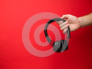 A young woman holds a pair of black wireless headphones in her hand. Hand of a girl with headphones on a red background