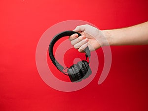 A young woman holds a pair of black wireless headphones in her hand. Hand of a girl with headphones on a red background