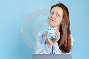 A young woman holds a piggy bank sitting with both hands and looks at the camera, with a laptop on her lap on a blue background