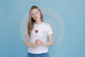 Young woman holds red heart on stick in white t-shirt on blue background.