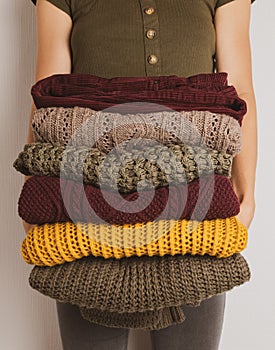 A young woman holds a stack of warm clothes in autumn colors. Sweaters in yellow, dark green, brown