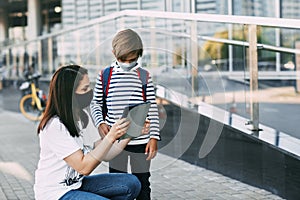 A young woman holds a tablet and shows her son the route. Mother and boy with a backpack in masks on the way to school, space for