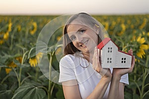 Young woman holds toy house on palms on field of sunflowers background. eco-friendly homes. Green houses