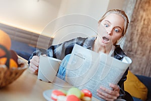 Young woman at home in the kitchen drinking tea reading schoked by news