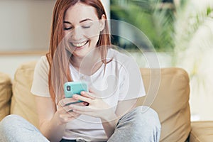 Young woman at home receiving positive emotions winning and good news while reading phone while sitting on sofa