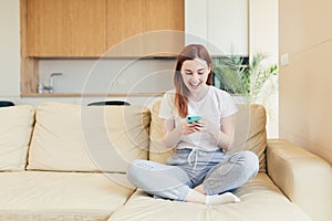 Young woman at home receiving positive emotions winning and good news while reading phone while sitting on sofa