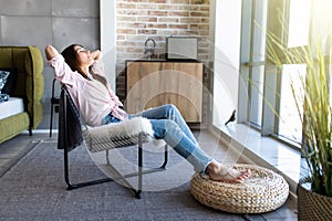 Young woman at home sitting on modern chair in front of window relaxing in her living room