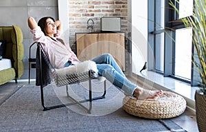 Young woman at home sitting on modern chair in front of window relaxing in her living room