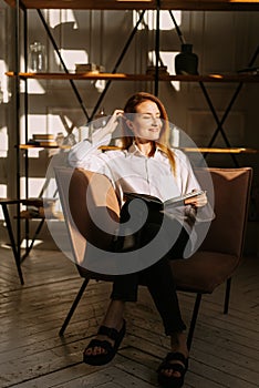 Young woman at home sitting on modern chair relaxing in her living room reading book. Eco modern style interior concept