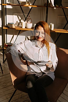 Young woman at home sitting on modern chair relaxing in her living room reading book. Eco modern style interior concept