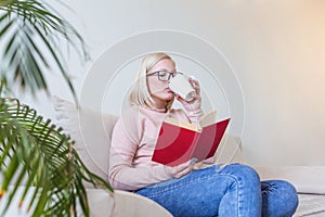 Young woman at home sitting on modern sofa relaxing in her living room reading book and drinking coffee or tea. White cozy bed and