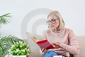 Young woman at home sitting on modern sofa relaxing in her living room reading book and drinking coffee or tea. White cozy bed and