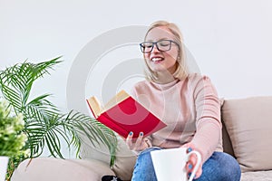 Young woman at home sitting on modern sofa relaxing in her living room reading book and drinking coffee or tea. White cozy bed and