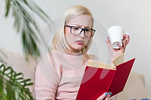 Young woman at home sitting on modern sofa relaxing in her living room reading book and drinking coffee or tea. White cozy bed and