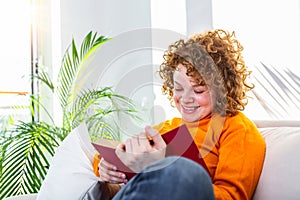 Young woman at home sitting on modern sofa relaxing in her living room reading book . White cozy bed and a beautiful girl, reading