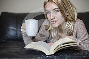 Young woman at home sitting near window relaxing in her living room reading book and drinking