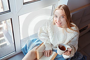 Young woman at home sitting on window sill winter concept drinking tea reading book