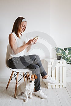 Young woman at home using mobile phone, cute small dog besides. work from home concept