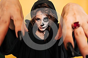A young woman in a hood with scary makeup. on a yellow isolated background. She& x27;s terrifying with her hands. Concert