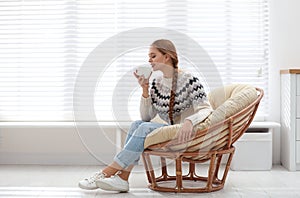 Young woman with hot drink in papasan chair near window