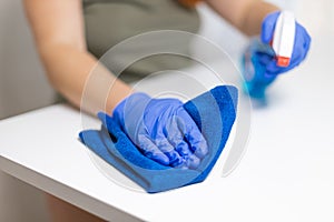 Young woman housekeeper is cleaning, wiping down table surface with blue gloves, wet rag, spraying bottle cleaner. COVID