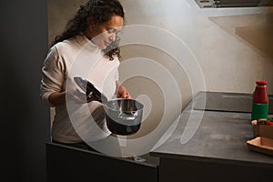 Young woman housewife takes out a stainless steel pan from the kitchen cabinet, standing at kitchen countertop