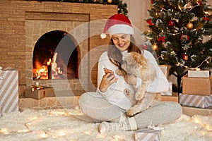 Young woman hugging Pekingese dog at christmas time, holding phone in hands and looking at device`s screen with smile, female wit