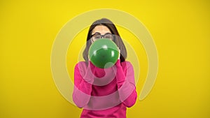 A young woman inflates a green balloon with her mouth on a yellow background. Girl in a pink turtleneck and glasses.