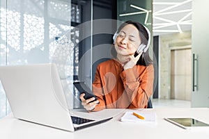 Young woman inside office at workplace listening to relaxing soothing music, Asian businesswoman in headphones using app