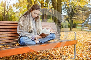 Young woman with interest reads book in autumn park. Girl sitting on bench on background of fallen yellow leaves