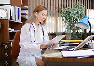 Young woman internist studies the patient's outpatient card, typing the treatment appointment on the computer in the