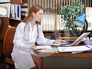 Young woman internist studies the patient's outpatient card, typing the treatment appointment on the computer in the
