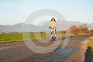 Mladá žena v bunde jazdí na bicykli po poľnej ceste, popoludňajšie slnko svieti na vrch Kriváň - slovenský symbol na pozadí