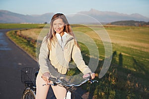 Young woman in jacket standing with a bicycle on asphalt country road, afternoon sun shines on fields meadows and mountains