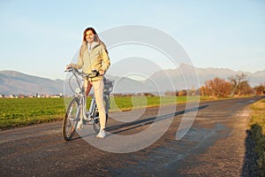 Young woman in jacket standing with a bicycle over country road, afternoon sun shines at mount Krivan peak - Slovak symbol -