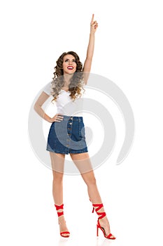 Young Woman In Jeans Mini Skirt And Red High Heels Is Pointing Over Head And Looking Up