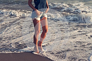 Young woman in jeans shorts walking through sea water at sunset sandy beach summer