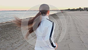 Young woman is jogging on the sand beach by the sea at sunrise in autumn, back view.