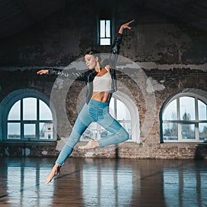 Young woman jumping in casual style - jeans and leather jacket doing ballet in old studio. Attractive ballerina