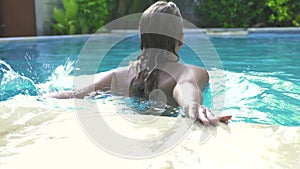 Young woman jumping in swimming pool on summer resort. Attractive girl swimming in blue water outdoor pool in resort