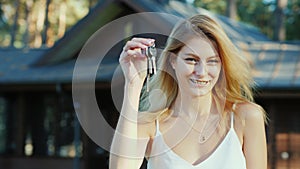 Young woman with keys from home. Smiling, looking at the camera. New wooden cottage in the background
