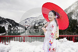 A young woman in a kimono, holding a red umbrella, stands on the bridge with snow and a boat. Toyama is located in Japan