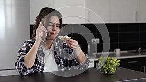 Young woman in kitchen during quarantine. Enjoying eating sandwich with cheese and phone conversation. Emotionally