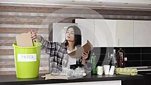 Young woman in kitchen during quarantine. Girl sorting paper from other trash and out it into green bucket. Zero waste