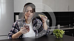 Young woman in kitchen during quarantine. Sit at table and eat sandwich with cheese. Chatting on smartphone. Social