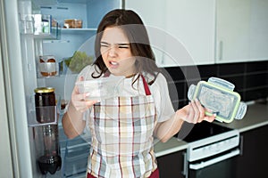 Young woman on kitchen during quarantine. Stand in front opened fridge with food tray with bad smell. Disgution.