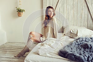 Young woman in knitted cardigan and warm socks wake up in the morning in cozy scandinavian bedroom and sitting on bed with white b