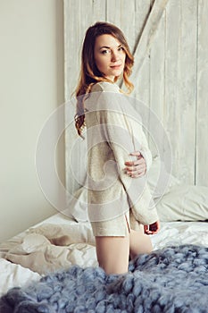 Young woman in knitted cardigan and warm socks wake up in the morning in cozy scandinavian bedroom and sitting on bed with white b