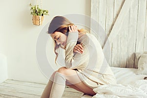 Young woman in knitted cardigan and warm socks wake up in the morning in cozy scandinavian bedroom and sitting on bed with white b